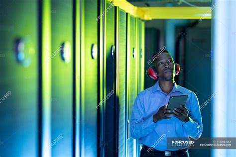 African american male computer technician wearing headphones using tablet working in server room ...