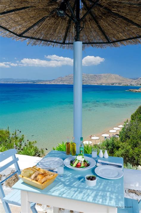 an outdoor dining area overlooking the ocean and beach with blue chairs, table and umbrella