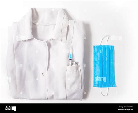 Flat lay medical gown with vaccine bottle and syringe in pocket and protective mask on white ...