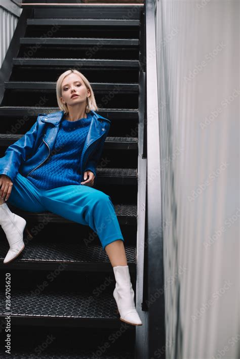 Blonde Woman Wearing Blue Leather Jacket And Heels Sitting On Stairs And Looking At Camera Stock