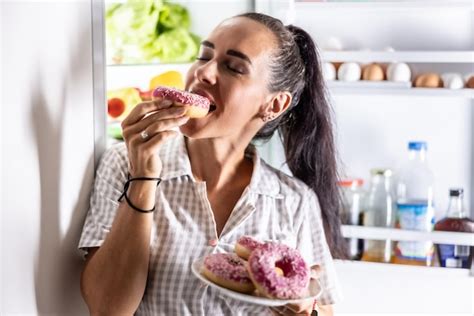 Premium Photo Hungry Brunette In Pajamas Enjoys Sweet Donuts Late At Night By The Open
