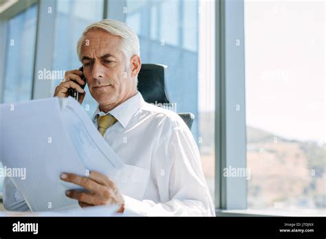 Shot Of Mature Businessman Sitting At His Desk Reading A Document And Talking On Mobile Phone
