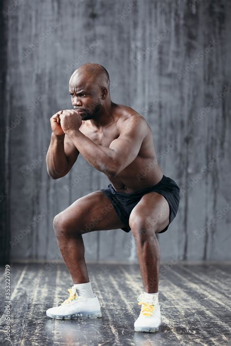 Strong And Muscular Dark Skinned Man With A Naked Torso Does Squats While Holding Hands In His