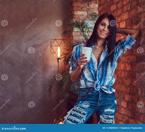 A Brunette In A Flannel Shirt And Jeans Holds A Cup Of Coffee Posing Against A Brick Wall Stock