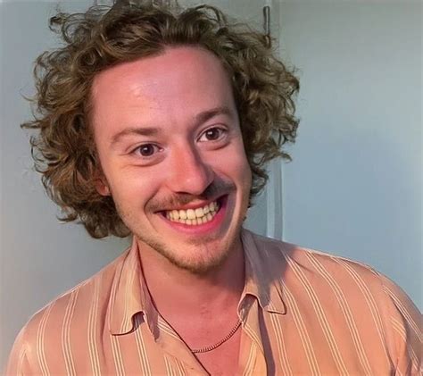 A Man With Curly Hair And A Mustache Smiles At The Camera While Wearing