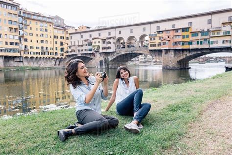 Lesbian Couple Sitting On Arno River Bank In Front Of Ponte Vecchio Holding Digital Camera