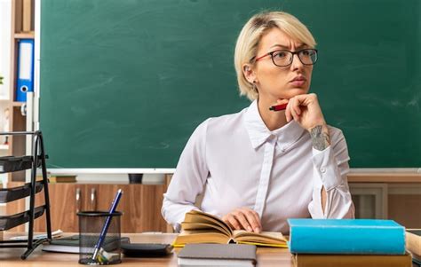 Jeune Enseignante Blonde Stricte Portant Des Lunettes Assise Au Bureau Avec Des Outils Scolaires