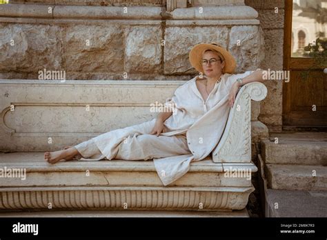 A Beautiful Mature Woman Sits On A Stone Bench In A Hat And Rests Stock