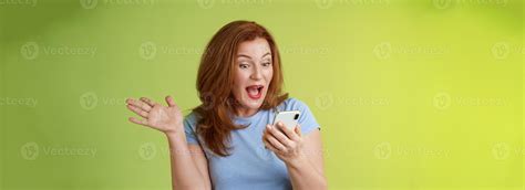 Surprised glad middle-aged pretty redhead woman look smartphone raise