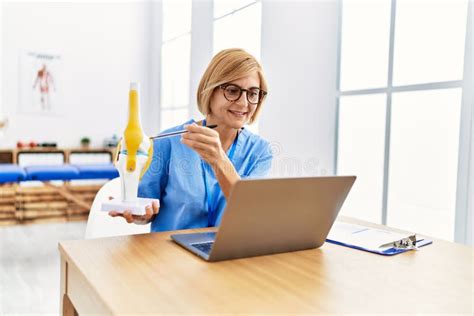 Middle Age Blonde Woman Wearing Physio Therapy Uniform Holding Anatomical Model Of Knee At