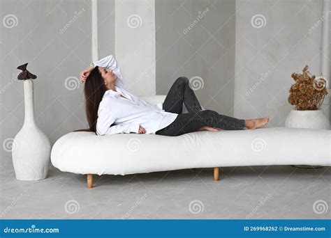 Slender Beautiful Brunette Girl In White Shirt And Black Jeans Resting On A White Sofa Stock