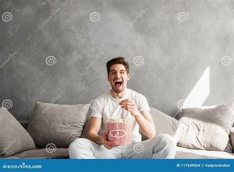 Joyous Young Man Sitting On Sofa In Living Room And Laughing While