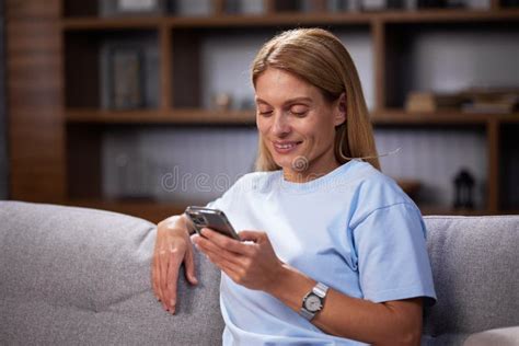 Blonde Girl Sitting On The Couch Looks Into The Smartphone Chatting Relaxing At Home Smiling