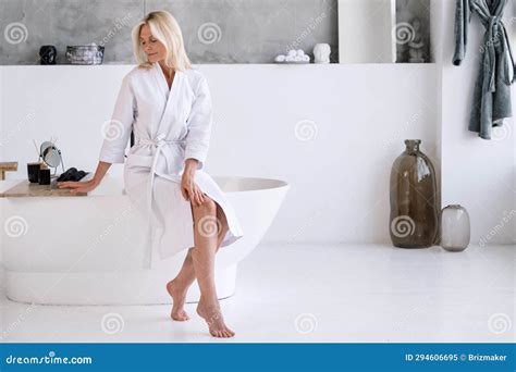 Mature Woman In White Bathrobe Sitting On Edge Of Bath Tub In Modern Bathroom Stock Image