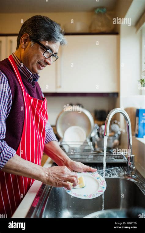 Mature Man Is Doing The Dishes In The Kitchen Of His Home He Is Wearing An Apron While Washing