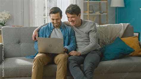 Adorable Male Gay Couple Spend Time At Home They Sit On A Sofa And Use The Laptop They Watch