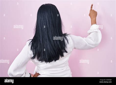 Mature Hispanic Woman Standing Over Pink Background Posing Backwards Pointing Ahead With Finger