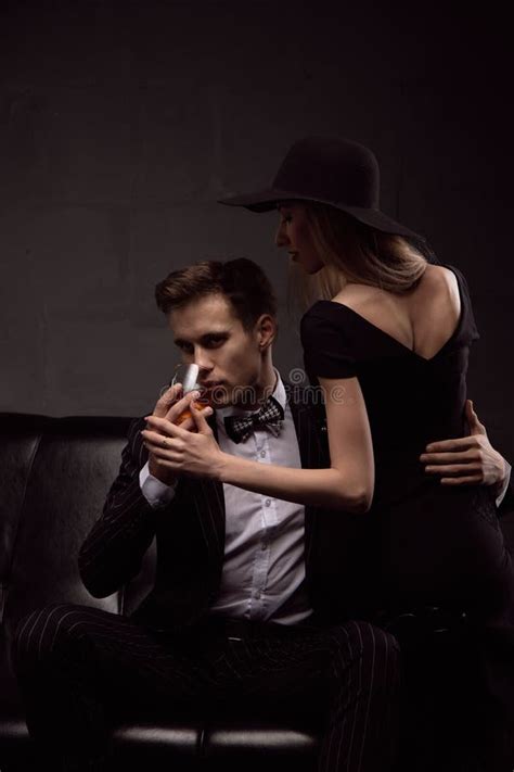 Rich Handsome Man Drinks Whiskey With A Blonde Mistress In The Evening In Low Light Stock Photo