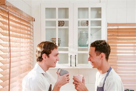 Happy And Lovely Caucasian Gay Couple Wearing Apron Drinking Coffee Or Tea While Looking At The