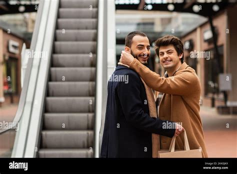Happy Gay Man In Beige Coat Hugging Neck Of Bearded Boyfriend Holding