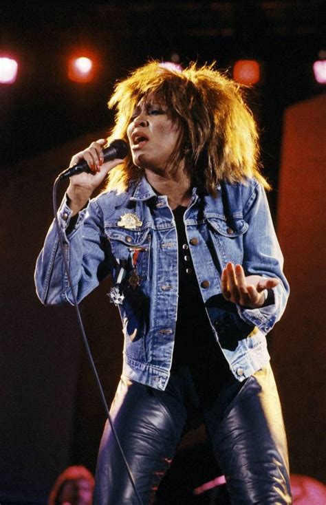 a woman in leather pants and denim jacket singing into a microphone with an audience behind her