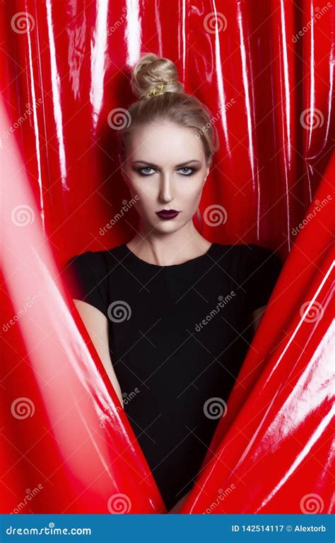 Beautiful Slender Blonde Girl Dressed In A Black Fitting Dress On A Background Of Red Latex
