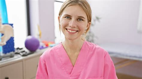 Attractive Blonde Woman With Blue Eyes Wearing A Pink Medical Scrub Smiles In An Indoor Rehab