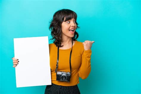Jovem fotógrafa latina isolada em fundo azul segurando um cartaz vazio e apontando o lado Foto