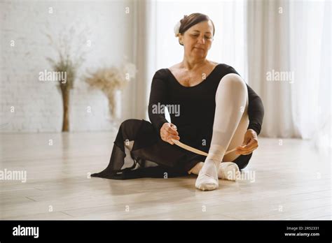 Full Body Of Mature Female Dancer In Black Dress Sitting On Floor And Putting On Ballet Pointe