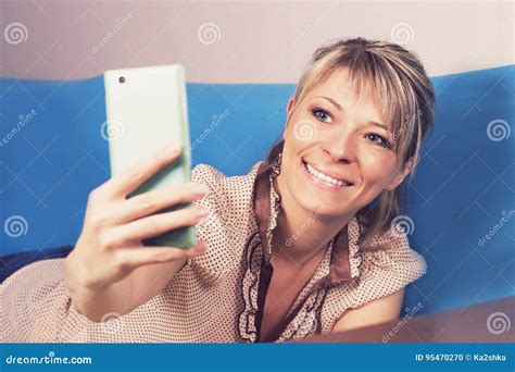 Fille Blonde Mignonne Sur Le Sofa Elle Regarde Son Smartphone Et Fait Un Selfie Photo Stock