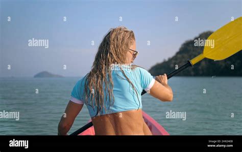 Long Curly Haired Blonde Woman In Swimsuit Rows Plastic Kayak Along Azure Ocean Water At Exotic