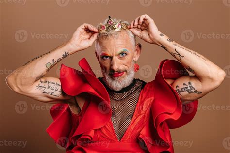 Portrait Of Confident Mature Gay Man In Beautiful Red Dress Wearing Crown Against Brown