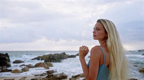 Blonde Woman In Blue Swimsuit Stands Serene On Rocky Seashore Ocean Waves Swell Tranquil Beach