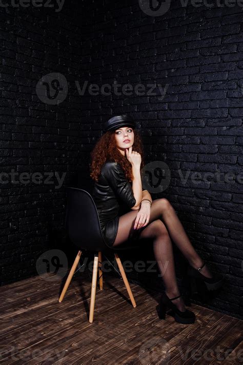 Brunette Curly Haired Long Legs Girl In Black Leather Dress And Cap Posed At Studio On Chair