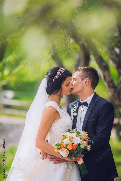 Elegant Stylish Groom With His Happy Gorgeous Brunette Bride On The Background Of Trees In The