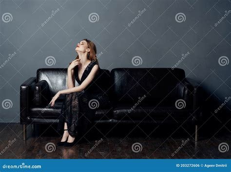 Woman In Black Dress Sits On Leather Sofa Indoors Evening Makeup Naked Shoulders Model Stock