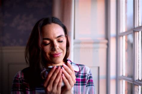 Woman With Closed Eyes At Home With Hot Drink Standing Looking Out Of Window With Evening Light