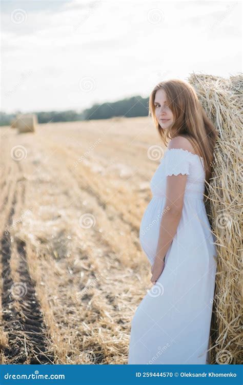 Beautiful Pregnant Brunette In A White Dress Near The Haystacks In The Field At Sunset Stock