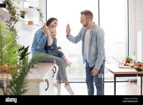 Beautiful Brunette Woman Sitting On Countertop With Baby On Laps While Handsome Man Standing In