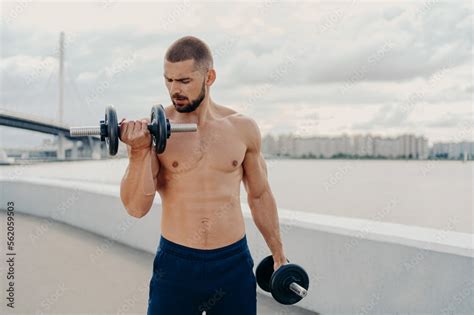 Muscular Guy With Naked Torso Exercises With Barbells Stands Shirtless Trains Muscles Outdoor