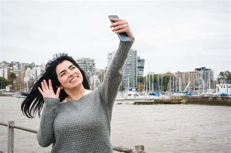 Joven turista latina de pie en el muelle tomando una selfie con su ciudad de teléfono en el