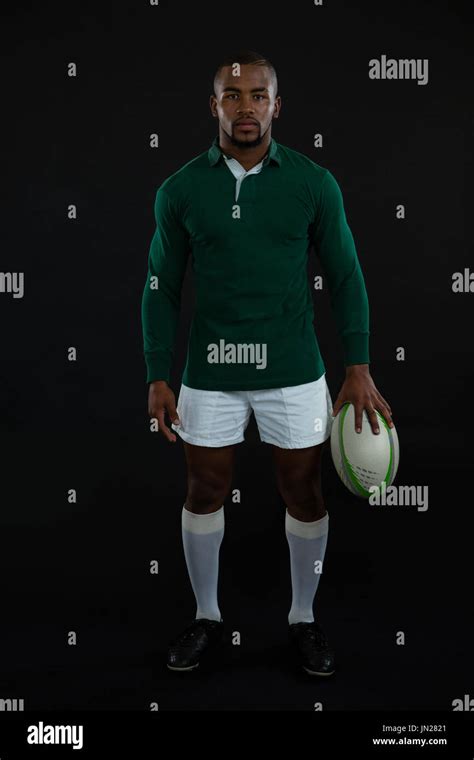 Portrait Of Male Rugby Player Holding Ball While Standing Against Black