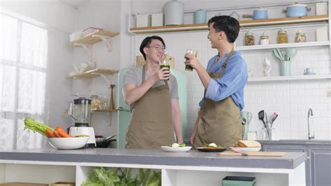 joven pareja gay sonriente haciendo jugo saludable con máquina de jugo en la cocina en casa