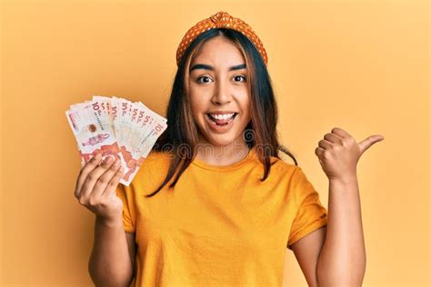 Jovem Latina Segurando Pesos Colombianos Apontando O Polegar Para O Lado Sorrindo Feliz A