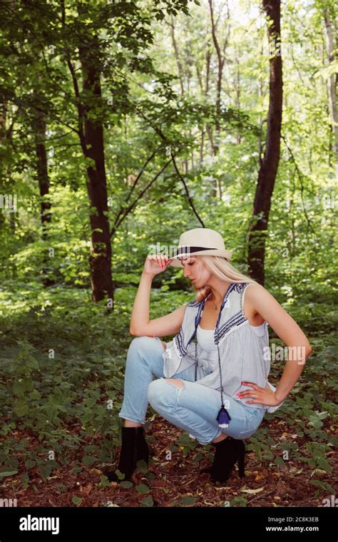 Blonde Girl Wearing A Hat Blue Jeans And Boots With High Heels Squats In A Park Or Forest With