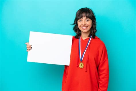 Joven latina con medallas aisladas de fondo azul sosteniendo un cartel vacío con expresión feliz