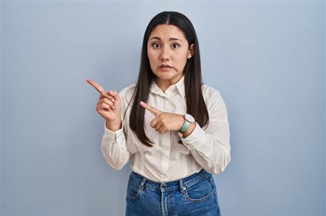 Joven latina parada sobre fondo azul señalando a un lado preocupada y nerviosa con ambas manos