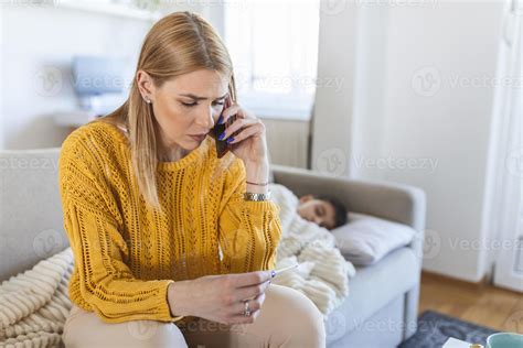 Concerned Caucasian Blonde Mother On Phone And Reading Thermometer While Her Kid Son Lies Sick
