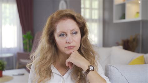 Thoughtful And Unhappy Mature Woman Woman Sitting Alone On Sofa At Home Looks Around And Is