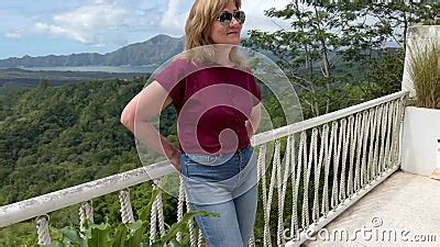 A Blonde Adult Woman Standing On A Terrace With White Rail With Dramatic Sight Of Volcano